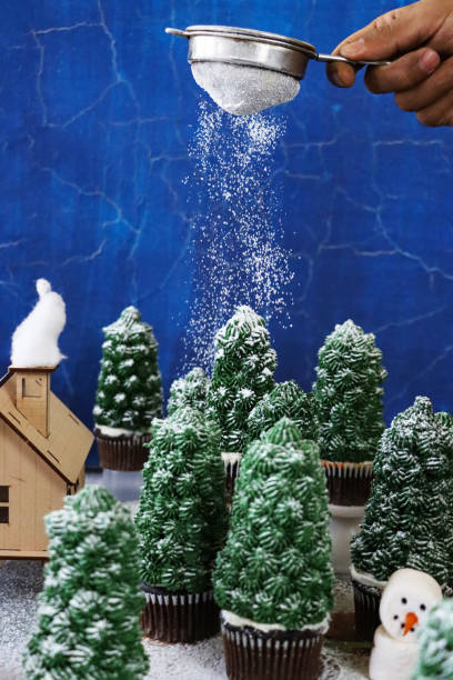istock Image of unrecognisable person sifting icing sugar snow over night conifer forest scene display of batch homemade, Christmas tree design chocolate cupcakes in paper cake cases, model house with marshmallow snowman 1349493485
