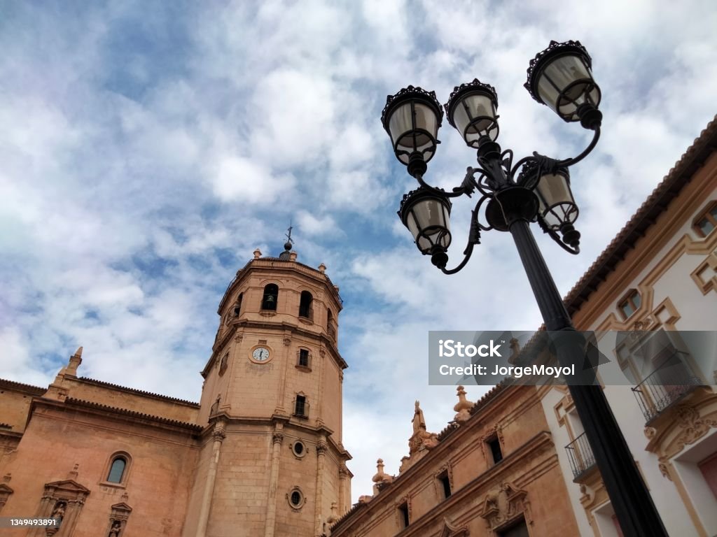 Church bell tower and lamppost View of part of the south façade of the church of San Patricio with tower and bell tower, XVI-XVIII centuries, in the Plaza de España of Lorca, Murcia, Spain and Fernando VII type lamppost on one side. Building Exterior Stock Photo