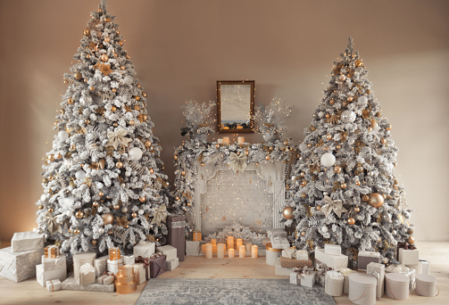 Hall room interior with a fireplace with candles and a two decorated Christmas trees with gifts. Large designer snow tree with lights and balls in beige, brown, gold and silver colors.