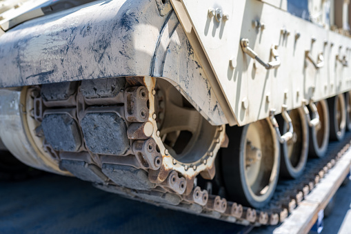 A close up on an old abandoned military truck, tank, or transporter made out of metal with rusty tracks attached to wheels seen on a dirt path located next to some logs, wood, spare parts, and forest