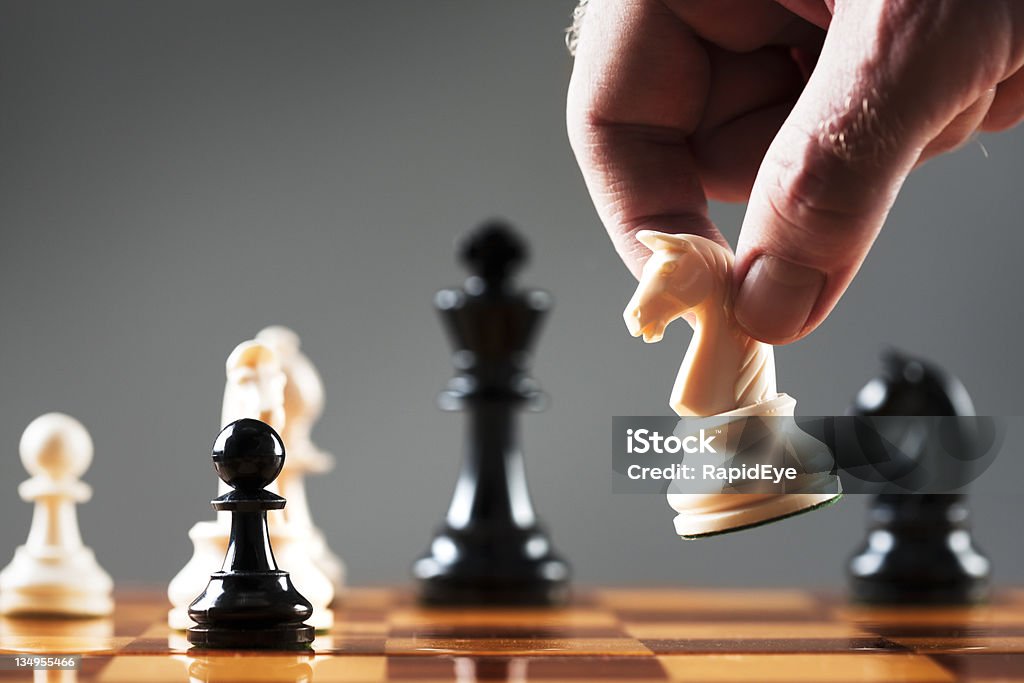 Man's hand moves white knight into position on chessboard A man's hand takes a white chess knight and prepares to make his move. White knight symbolism, meaning corporate rescue from a takeover by a friendly investor. Shot with Canon EOS 1Ds Mark III. Chess Stock Photo