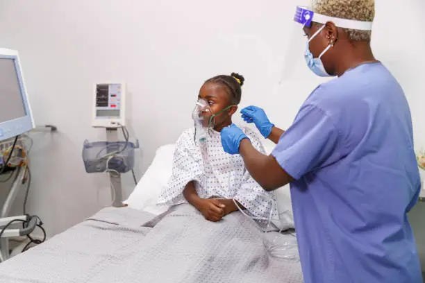 A preadolescent black girl who is hospitalized while sick with Coronavirus sits in her hospital bed as a nurse helps her use a medical ventilator for breathing assistance.