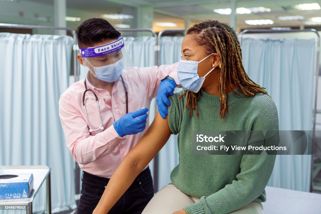 Black woman receiving COVID-19 vaccination injection A young black woman wearing a protective face mask sits in a medical clinic as her male doctor of Asian descent prepares to give her a COVID-19 vaccination injection in the upper arm. COVID-19 Vaccine Stock Photo