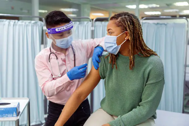 A young black woman wearing a protective face mask sits in a medical clinic as her male doctor of Asian descent prepares to give her a COVID-19 vaccination injection in the upper arm.