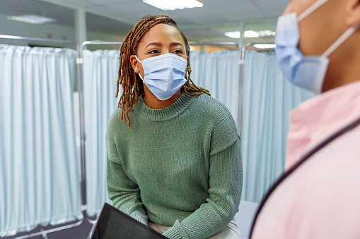 A black woman wearing casual clothing sits on a hospital bed and speaks with her doctor. The woman is being discharged after a hospital stay and she is reviewing with her doctor test results and a treatment plan which are displayed on a tablet computer. Both individuals are wearing protective face masks.