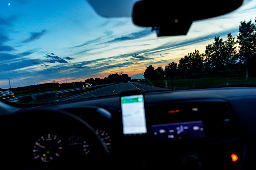 Speeding car driver point of view looking through the windshield above the dashboard and a blurred, generic smart phone device screen navigation map. Dramatic sky dusky night multiple lane expressway just after sunset with a bright crescent moon shining in the upper left corner of the image.