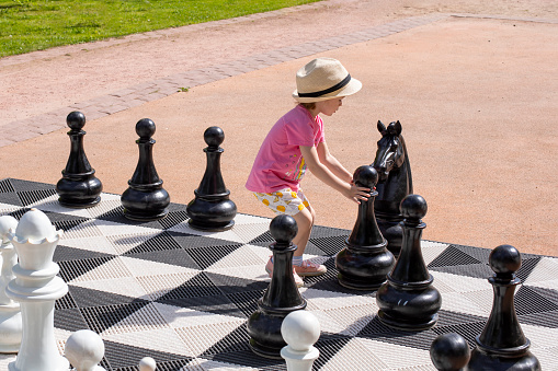 Little Caucasian girl playing giant chess in the park, horizontal photography. Girl making a pawn move