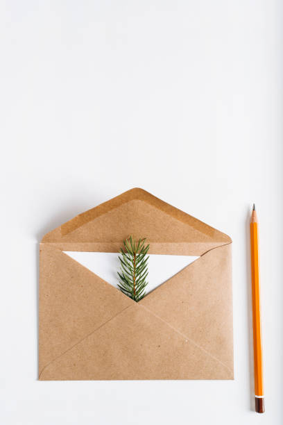 istock Christmas craft envelope with greeting card inside, with an invitation or xmas wish. Flat lay brown open envelope, paper card, pine twig and pencil on white table, copy space for text. Vertical photo 1349603531