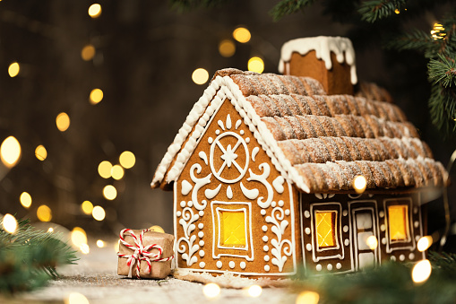 Gingerbread House with Christmas Gift over Shining Garland Lights Background. Xmas Holiday Present Art Decoration. Baked Sweet Ginger Cake with Icing over Defocused Bokeh