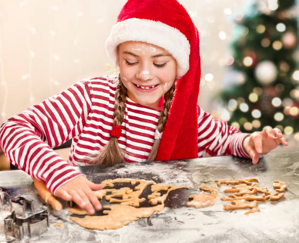 una chica con sombrero de papá noel hace galletas en la mesa - christmas child cookie table fotografías e imágenes de stock