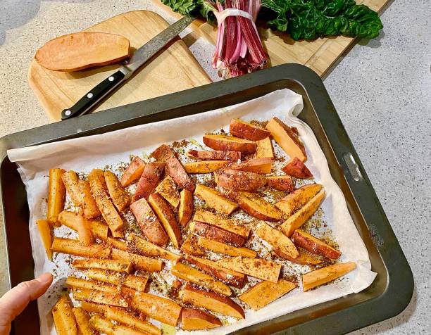 Home Made Sweet Potato Fries Horizontal close up tray of thick cut sweet potato fries chips on baking paper with herbs on kitchen bench held by hand ready for placing in oven sweet potato french fries stock pictures, royalty-free photos & images