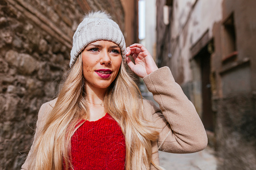 Portrait of young blond latin woman in stylish coat and woolen hat posing in old street. Argentinian woman touching her hat.