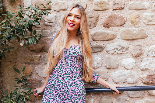 Blond latin smiling woman in stylish colorful dress leans against stone wall in the street. Cheerful beautiful argentinian woman looking at camera.