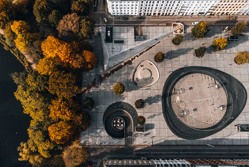 Aerial view of Israels Plads (Israels Square) and cars driving into underground parking garage in yellow autumn colours. Once a parking lot filled with cars, this public plaza has now been transformed into a modern square facilitating street sports and performance venue, a school yard and a thriving market. The design by Cobe Architects includes an underground parking garage in an attempt to create a sustainable urban environment favouring the citizens of Copenhagen. Image shot with drone.