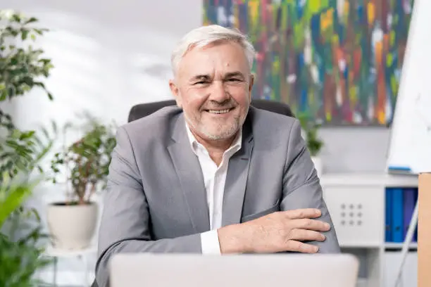 Close-up of face of smiling man in shirt jacket, guy with gray hair stubble in old age is sitting on comfortable chair in office of company is looking into camera friendly boss hands crossed on chest