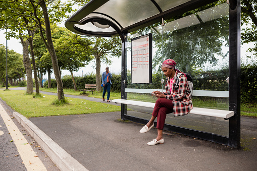 A businesswoman sitting at a bus stop using her mobile phone while waiting for a bus in Newcastle-upon-Tyne. There is a businessman walking in the background.