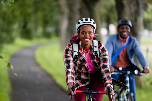A businessman and businesswoman wearing bike helmets, riding bikes through a public park on a footpath in Newcastle-upon-Tyne on their commute to work. The woman is riding ahead, looking at the camera and smiling while the man, who is out of focus, is following her and smiling.