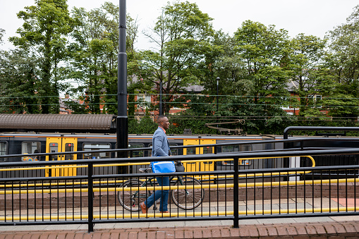 A businessman wearing casual businesswear and a satchel, walking up a ramp after getting off a train in Newcastle-Upon-Tyne. He is pushing his bike while he walks to work. The train is standing stationary in the station behind him, preparing to leave.