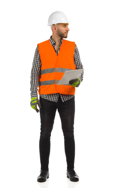 istock Engineer in orange reflective waistcoat and white helmet is standing with documents and looking at the side. 1349673116