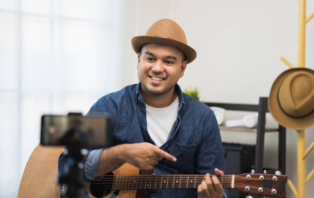 Young asian musician teaching the guitar class online live streaming with smartphone with student. Indian man playing acoustic guitar while singing a song and record by cell phone shaed online social joven músico asiático que enseña la clase de guitarra en línea transmisión en vivo con teléfono inteligente con el estudiante. hombre indio tocando la guitarra acústica mientras canta una canción y graba por teléfono celular shaed online social