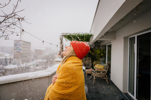 Photo of a young woman wrapped in a blanket on the terrace of her loft apartment on a cold snowy day.