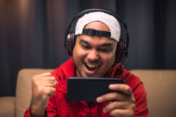 istock Playing video game on smartphone. Young asian handsome man sitting on sofa holding cellphone in living room. Happiness Streamer Indian man wearing headset playing game online in the darkroom. 1349688949