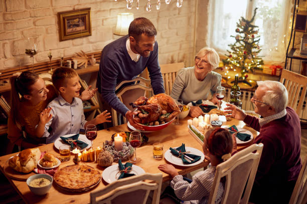 Happy man serving roast turkey for his family during Thanksgiving dinner in dining room. Happy extended family celebrating Thanksgiving and having traditional meal at dining table. Focus is on man serving roast turkey. merry christmas family stock pictures, royalty-free photos & images
