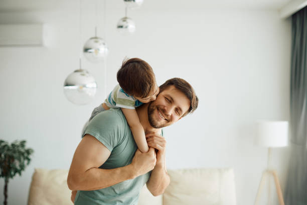 Happy father enjoying in spending time with his small son at home. Little boy kissing his father who is piggybacking him while spending time together at home. parenting stock pictures, royalty-free photos & images