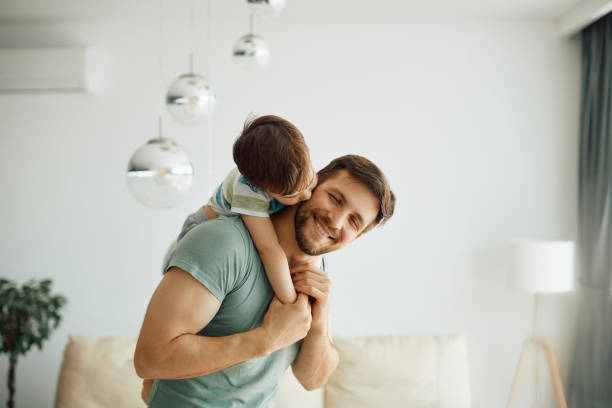 Happy father enjoying in spending time with his small son at home. Little boy kissing his father who is piggybacking him while spending time together at home. father and son stock pictures, royalty-free photos & images