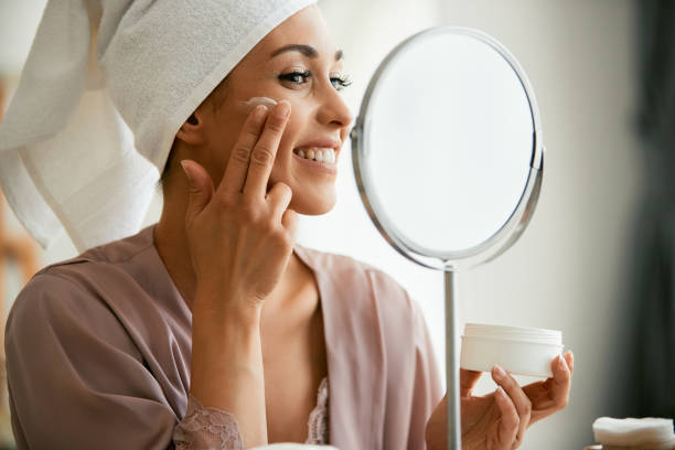istock Happy woman applying face cream while looking her self in a mirror. 1349701502