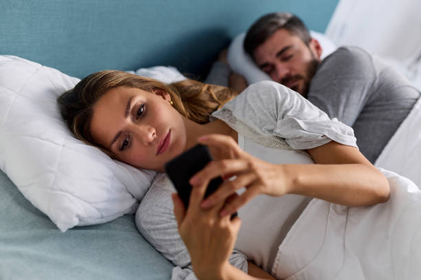mujer joven usando el teléfono móvil y revisando mensajes en la cama mientras su esposo está dormido - deshonestidad fotos fotografías e imágenes de stock