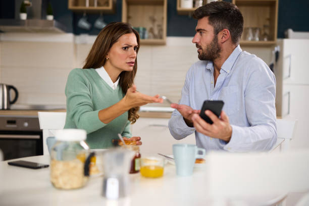 Young woman looking irritated while her husband uses his phone during breakfast at home stock photo