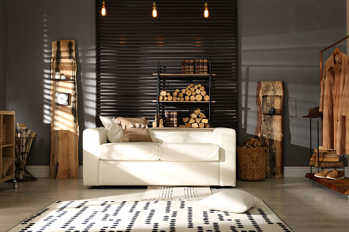 Shelving unit with stacked firewood and comfortable sofa in stylish room interior