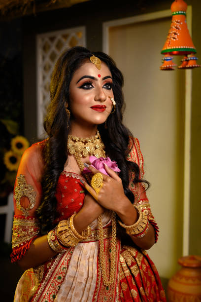 Portrait of pretty young Indian girl wearing traditional saree, gold jewellery and bangles holding lotus flower in her hands in studio lighting indoor. Indian culture, occasion, religion and fashion. stock photo