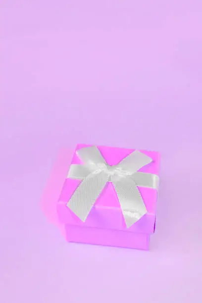 Purple-lilac gift box with a white bow on a vertical neon background. Minimalistic vertical photo of a gift box.
