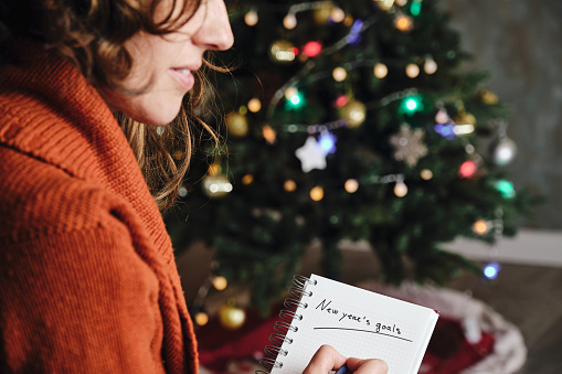 Mujer caucásica sonriendo con suéter naranja de perfil con cuaderno de metas de Año Nuevo en mano con árbol de Navidad desenfocado en el fondo photo