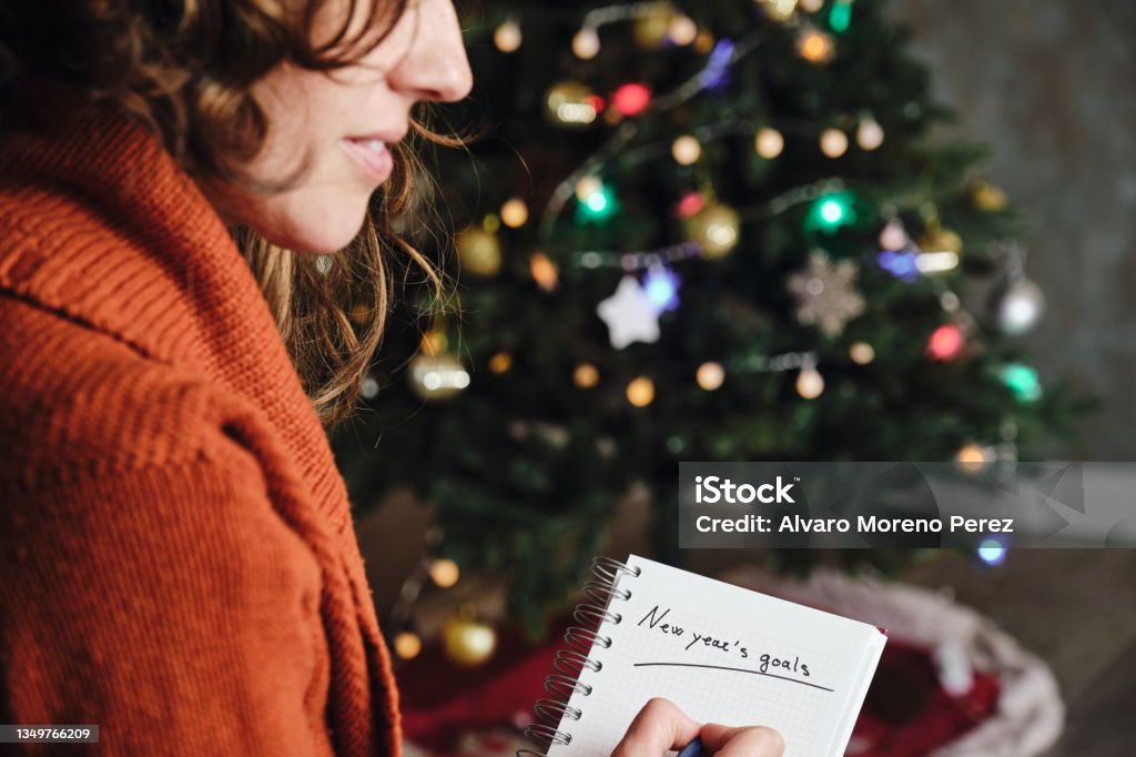 Caucasian woman smiling with orange sweater in profile with new year's goals notebook in hand with unfocused christmas tree in the background New Year Resolution Stock Photo