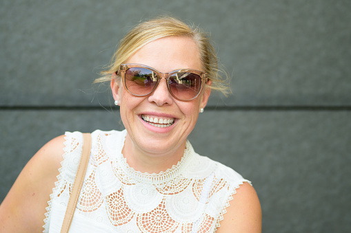Close up portrait of stylish blond woman wearing sunglasses standing against a grey wall