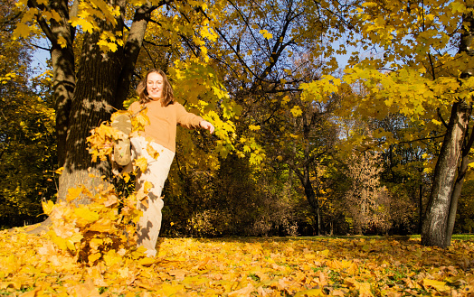 Defocused young laughing woman kikking dry autumn leaves with her foot in the forest on sunny day. Happiness.