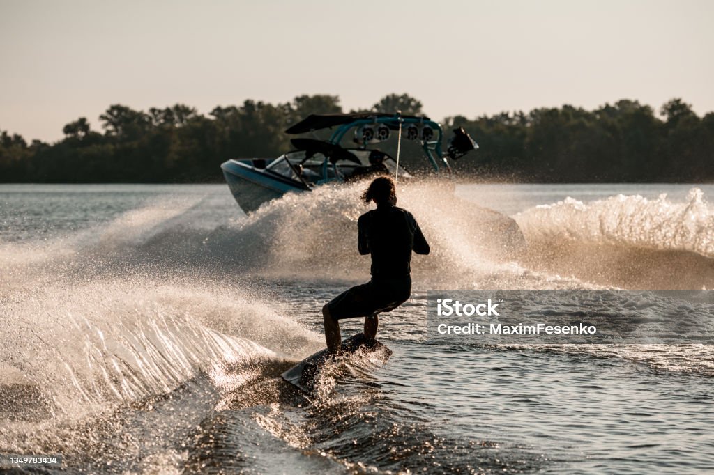 Rear view of active man riding wakeboard behind motor boat on splashing river waves. Active and extreme sports Rear view of athletic guy holding rope and energetically riding wakeboard behind motor boat on splashing river waves. Active and extreme sports Wakeboarding Stock Photo