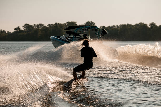 Rear view of active man riding wakeboard behind motor boat on splashing river waves. Active and extreme sports stock photo