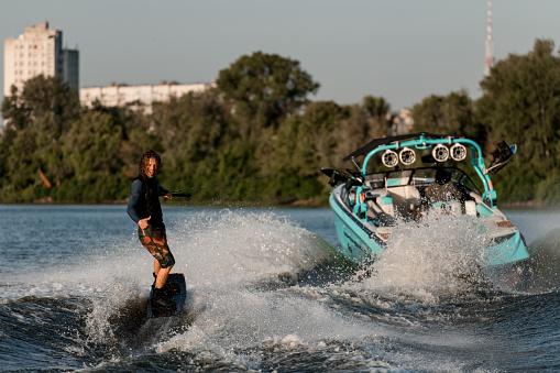 Bright motor boat pulls an active wet man riding a wakeboard down the river and showing surfer gesture. Summertime watersports activity