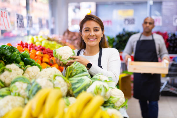 Smiling supermarket workers in fruit and vegetables section Latin saleswoman in  black apron and  her assistant with box of vegetables in  background merchandiser stock pictures, royalty-free photos & images