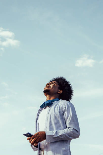 młody afro mężczyzna z telefonem komórkowym patrzący w niebo. tło nieba. pionowa przestrzeń kopiowania. - young men looking up thinking looking zdjęcia i obrazy z banku zdjęć