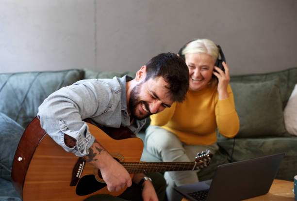 A young man is playing the guitar while his mother listens to him. She has the headphones on her head. A talented young man is sitting and playing the guitar. His mother is sitting on the sofa bed with headphones on and listening to music he plays. The young man decided to surprise his mother with a song he wrote by himself. father and son guitar stock pictures, royalty-free photos & images