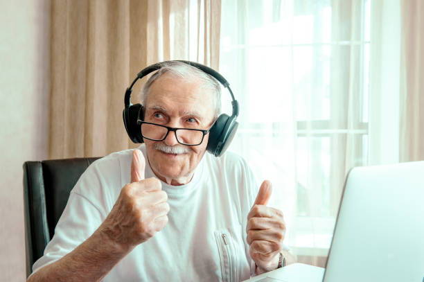 portrait of an elderly man with headphones. Positive human emotions, facial expression. A smiling elderly man using a laptop at home, listening to his favorite music and looking for information on the Internet stock photo
