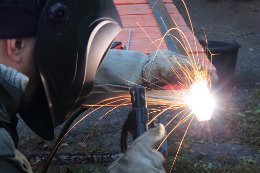 Welder wearing a protective mask and gloves while working. Welder craftsman makes a metal frame in the backyard.
