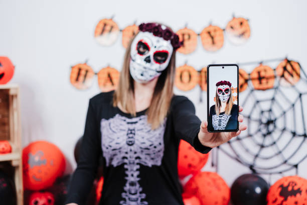 ハロウィーンのお祝いの間にメキシコのフェイスマスクを身に着けている女性。携帯電話で自分撮りを取る。頭にスケルトンの衣装と赤いバラのダイアデムを身に着けている女性。ハロウィ� - single flower flower autumn pumpkin ストックフォトと画像