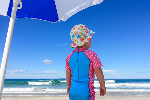 Child in sun hat, under a sun umbrella, looking out to sea, Omaha Beach, Auckland, New Zealand, summer holiday