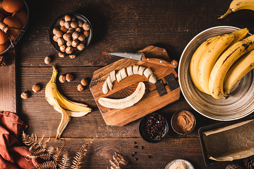 High angle view of a chopped banana on chopping board with hazelnuts, chocolate chips, brown sugar and cinnamon for making banana bread. Baking banana bread in the kitchen.